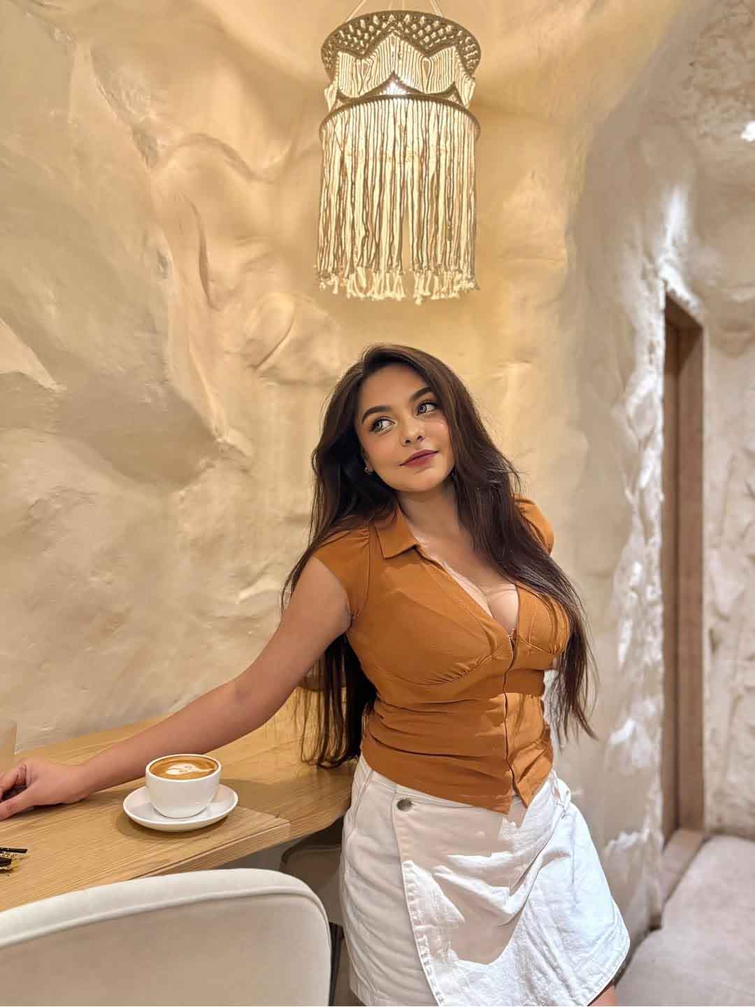 Anika Premium Escorts in Indore standing near a café counter, wearing a brown top and white skirt.