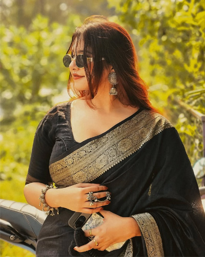 Avni Independent Escorts in Indore looking elegant in a black and gold saree, standing on a sunlit road surrounded by greenery.