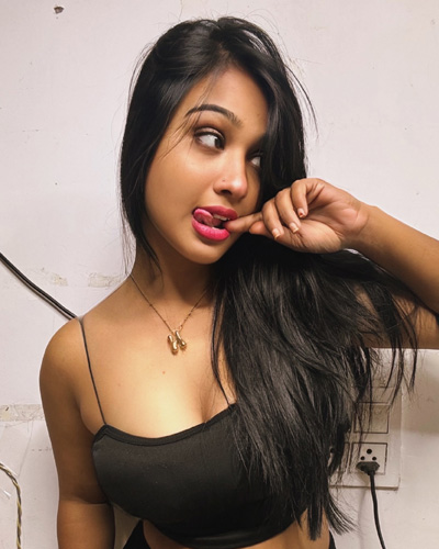 A woman with long black hair wearing a black sleeveless top, looking to the side while lightly touching her finger to her lips, standing indoors against a plain wall.