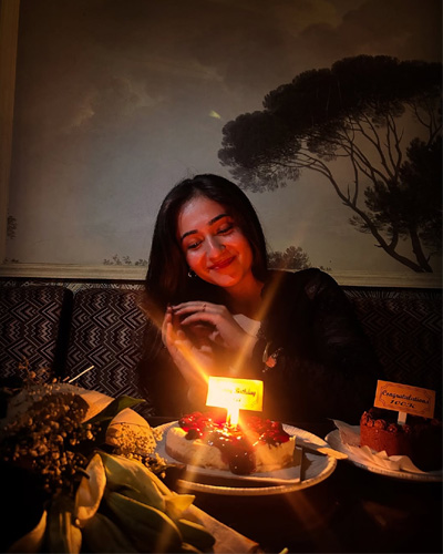 Riya Super Hot Escorts in Indore seated in a beautifully decorated restaurant, wearing a deep-neck black lace dress and posing gracefully with a drink placed in front