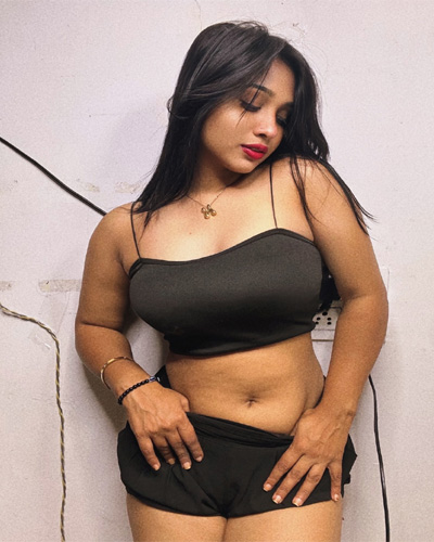 A woman with long black hair wearing a black sleeveless top and shorts, looking downward while standing indoors against a light-colored wall.